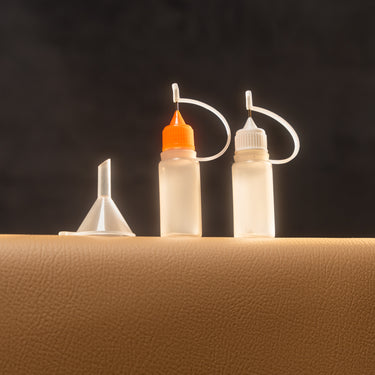 2 plastic paint dropper bottles with colored caps, used for applying small amounts of paint or finish, displayed on a brown leather surface in front of a dark background.