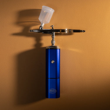 A cordless air brush used for applying paint or finishes, branded with the Maker's leather Supply logo, laid out on a light brown leather textured background.