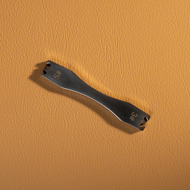 Metal zipper remover tool used for removing zippers on apparel and fashion goods, laid out on a light brown textured leather background.