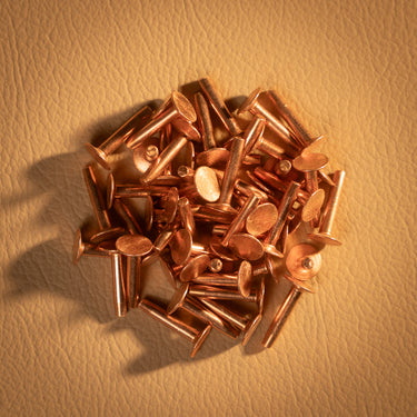 A pile of copper rivets used for leather crafting and other craft projects. Laid out on a brown textured leather background.