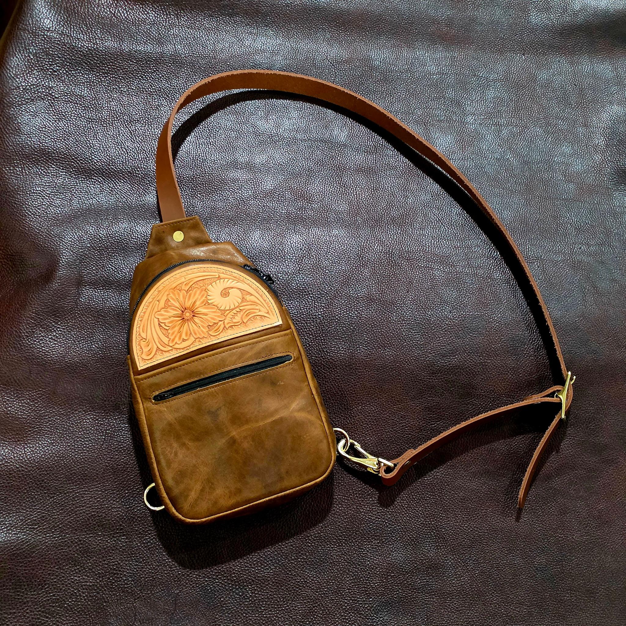 Leather sling bag with a floral tooled leather patch, zipper pocket, and adjustable strap.